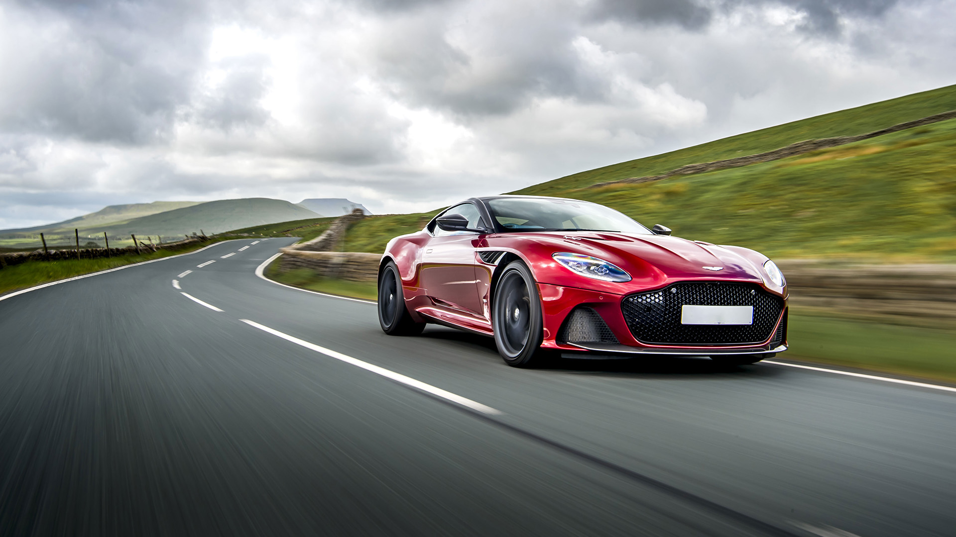 Red Aston Martin DBS driving fast on a country road with grey sky above.