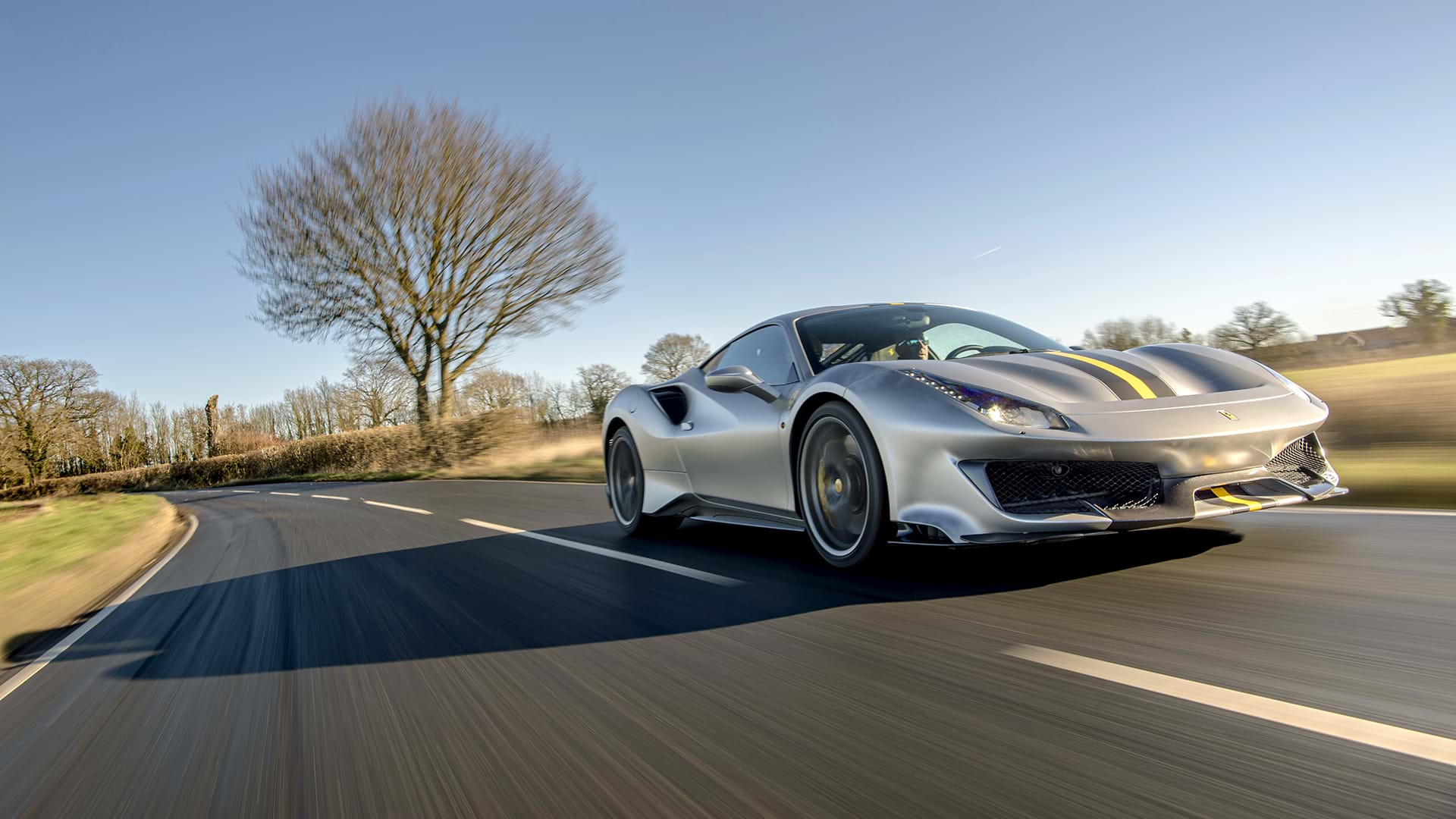 Sleek grey Ferrari 488 Pista with a bold racing stripe cruising through a scenic country landscape. 